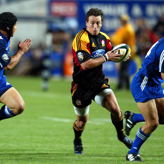 Chiefs rugby player runs with the ball against two defenders during a Super Rugby match, showing speed, focus, and determination — part of the proud Chiefs legacy celebrated by Ruakura Rugby Club.